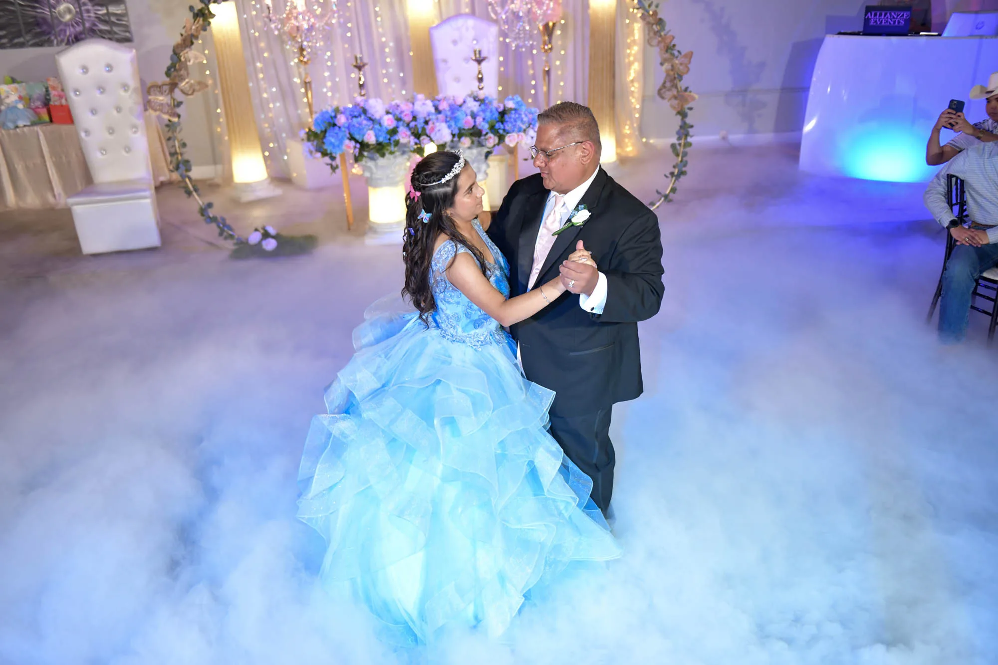 A father and daughter dancing at a quinceanera in the Moonlight Ballroom, surrounded by low fog and a lit floral backdrop